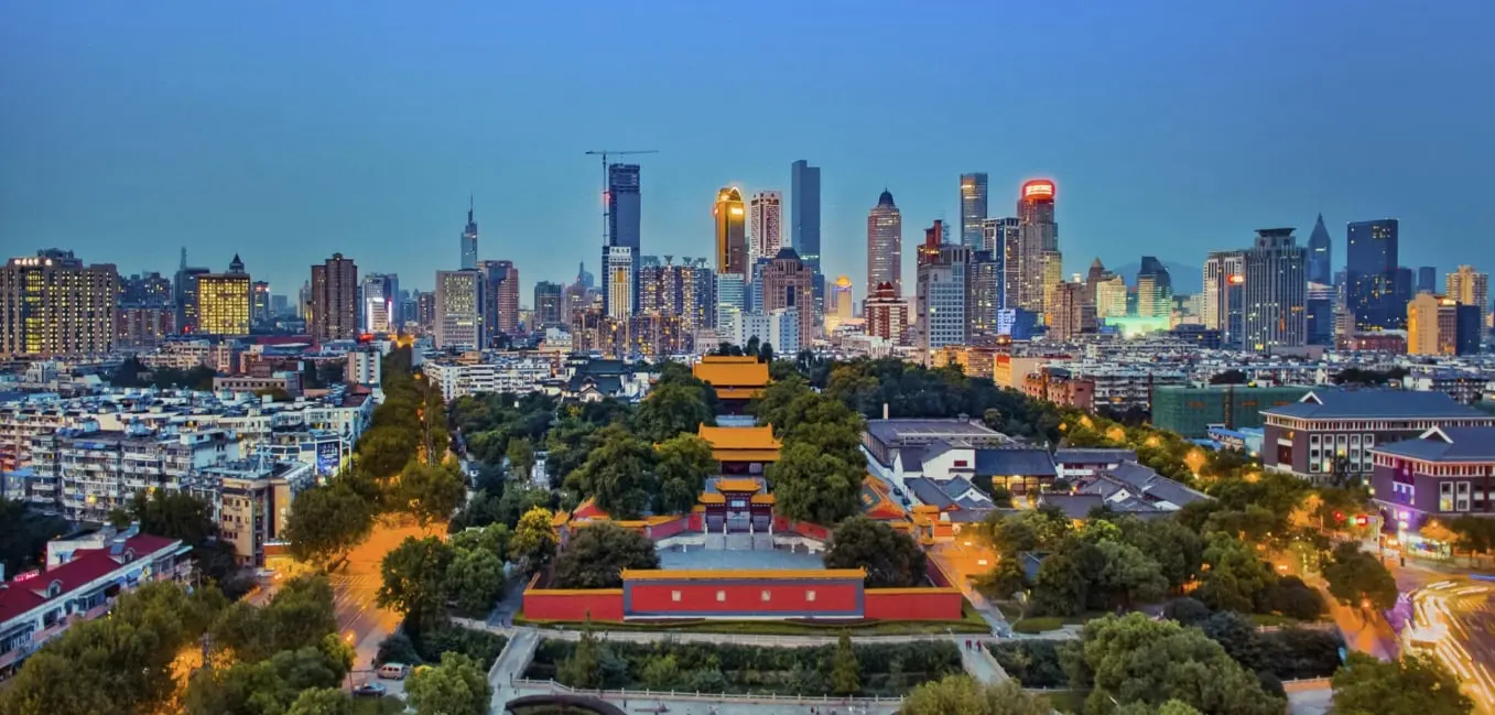 Vista espectacular de una ciudad al atardecer que combina elementos históricos y modernos. En primer plano se encuentra un templo tradicional rodeado de frondosos árboles, destacando su arquitectura clásica con tejados rojos y amarillos. Al fondo, imponentes rascacielos iluminan el horizonte, representando el desarrollo urbano. Una avenida arbolada divide la escena, mostrando el equilibrio entre la naturaleza y la urbanización. Perfecto ejemplo de armonía entre lo antiguo y lo moderno.