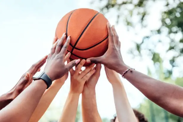 Imagen de varias manos levantadas intentando alcanzar un balón de baloncesto. Las manos pertenecen a personas de diferentes tonos de piel, reflejando diversidad e inclusión. El balón está sostenido en el aire mientras los jugadores se esfuerzan por tocarlo al mismo tiempo. El fondo muestra un entorno al aire libre con árboles y un cielo parcialmente visible, sugiriendo un partido o juego recreativo en un parque o cancha exterior. La imagen evoca una sensación de trabajo en equipo, competencia amistosa y espíritu deportivo.