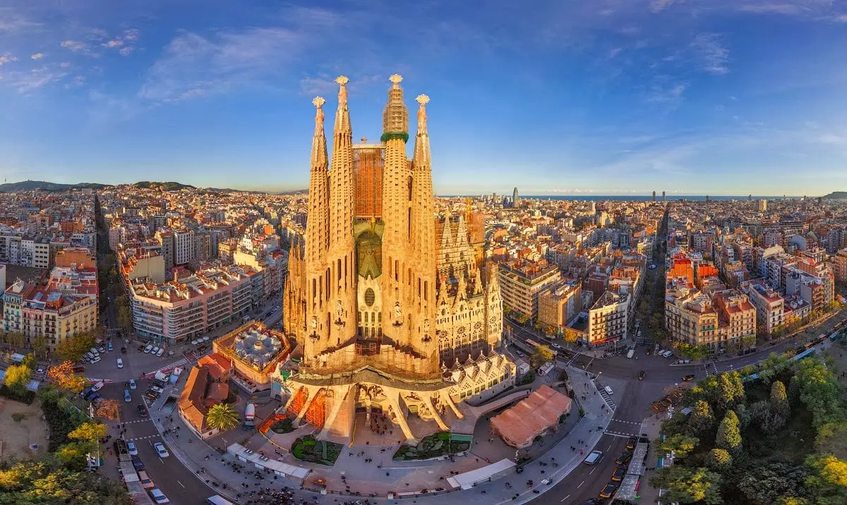 Impresionante vista aérea de la Sagrada Familia, el icónico templo diseñado por Antoni Gaudí en el corazón de Barcelona. Rodeada por un entramado urbano perfectamente simétrico, la basílica resalta con sus majestuosas torres bañadas por la cálida luz del atardecer. Al fondo, el mar Mediterráneo y el skyline de Barcelona completan esta escena única, capturando la mezcla de arte, arquitectura y modernidad de la ciudad.