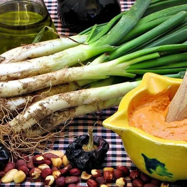 La imagen destaca los calçots con salsa romesco, un plato icónico de la gastronomía catalana. Los calçots, tiernos y asados, se combinan con la salsa romesco hecha de almendras, avellanas, pimientos y aceite de oliva. Este manjar tradicional es protagonista de las calçotadas, celebraciones típicas de invierno que reúnen a familias y amigos para disfrutar de la cocina catalana auténtica.