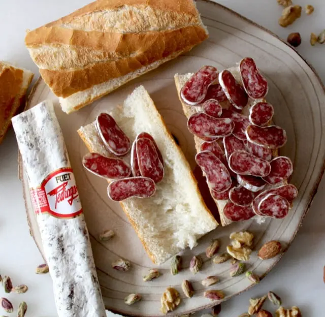 Esta imagen muestra un delicioso bocadillo de fuet, un embutido tradicional catalán. Acompañado de pan fresco, frutos secos como pistachos y nueces, el fuet destaca por su sabor único y textura suave. Este clásico de la gastronomía catalana es perfecto para aperitivos o meriendas, reflejando la riqueza culinaria y el gusto por los productos locales en Cataluña.