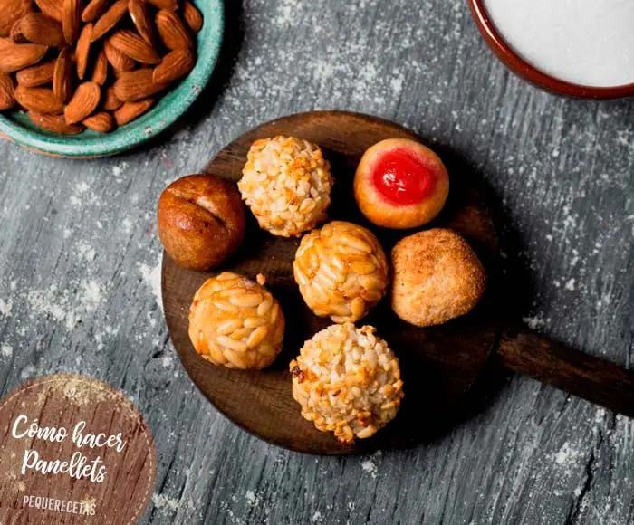 La imagen presenta panellets, los dulces tradicionales de Cataluña elaborados especialmente para la festividad de Todos los Santos. Estos pequeños bocados, hechos a base de almendra molida, azúcar y patata o boniato, se decoran con piñones, coco, cerezas o cacao. Los panellets son un símbolo de la repostería catalana, ideales para disfrutar con la familia en una ocasión especial.