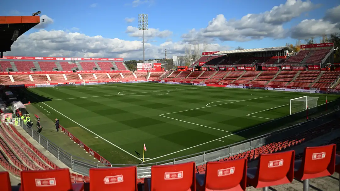 La imagen muestra el Estadi Montilivi, casa del Girona FC. El campo luce impecable con su césped perfectamente marcado, rodeado de gradas vacías en rojo vibrante, reflejando la identidad del club. Bajo un cielo azul con nubes dispersas, el estadio espera la emoción de un partido, listo para recibir a jugadores y aficionados. Un lugar lleno de historia futbolística.