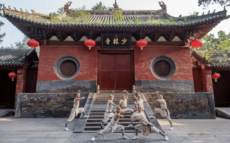 Monjes Shaolin realizando una exhibición de kung-fu frente al histórico Templo Shaolin en Dengfeng, China. La imagen captura la esencia de las artes marciales tradicionales en el icónico templo budista, rodeado de arquitectura antigua y decorado con faroles rojos chinos. Ideal para quienes buscan información sobre la cultura Shaolin, destinos turísticos en China, y el impacto del kung-fu en el patrimonio mundial.