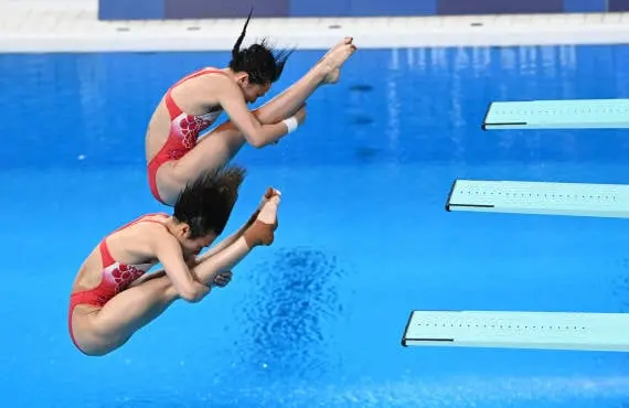 Atletas realizando clavados sincronizados desde un trampolín en una competición de deportes acuáticos. La imagen captura la precisión y coordinación de las deportistas mientras ejecutan acrobacias en el aire antes de entrar al agua. Ideal para contenido relacionado con los clavados sincronizados, deportes olímpicos, la técnica en las disciplinas acuáticas y el entrenamiento de alto rendimiento en natación.