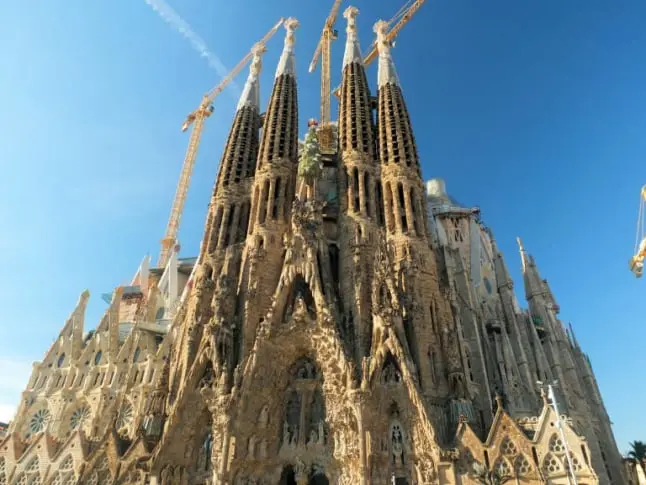 La imagen muestra la icónica Basílica de la Sagrada Familia en Barcelona, diseñada por Antoni Gaudí, destacada por su arquitectura modernista y su estilo único. La fotografía captura la fachada de la Natividad, una de las más elaboradas, con esculturas detalladas que representan escenas bíblicas y elementos de la naturaleza. Las torres imponentes, aún en construcción, se elevan hacia el cielo, rodeadas de grúas que simbolizan el proceso continuo de su creación. El diseño combina formas orgánicas, detalles geométricos y un simbolismo profundamente religioso, haciendo de la Sagrada Familia un emblema cultural y arquitectónico de Cataluña.