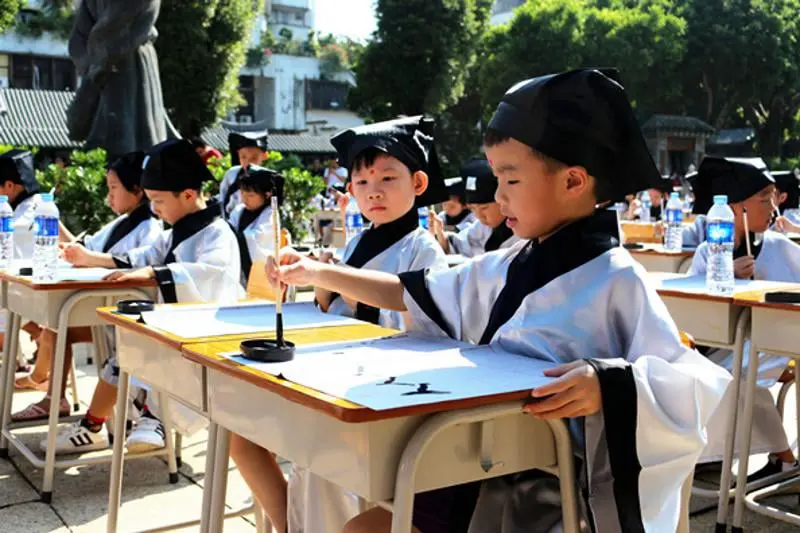 Niños chinos vestidos con trajes tradicionales practicando caligrafía en un ambiente cultural al aire libre. Esta escena resalta el valor de preservar las tradiciones ancestrales en la educación moderna. Ideal para destacar la importancia de la caligrafía como arte y disciplina en la formación de habilidades motoras, paciencia y conexión con la rica herencia cultural china.