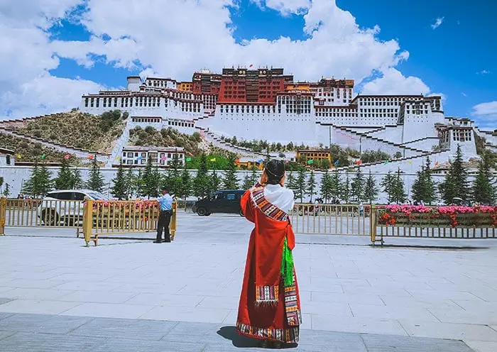 La imagen muestra el majestuoso Palacio de Potala en Lhasa, Tíbet, una joya arquitectónica y espiritual. Una visitante con traje tradicional tibetano resalta la riqueza cultural de la región. Este Patrimonio de la Humanidad por la UNESCO combina historia, religión y belleza, siendo un destino imprescindible para explorar la herencia cultural del Tíbet y disfrutar de paisajes únicos en las alturas.