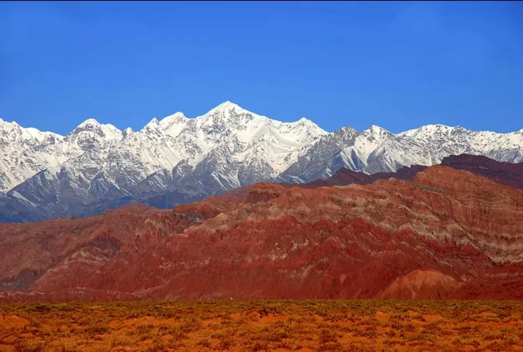 La imagen muestra el espectacular contraste de las Montañas Tian Shan en China, con picos nevados en el horizonte y formaciones rocosas rojizas en primer plano. Este paisaje único combina belleza natural y diversidad geológica, ofreciendo un destino ideal para aventureros y amantes de la naturaleza. Las Montañas Tian Shan son Patrimonio de la Humanidad por la UNESCO, perfectas para explorar la grandiosidad de China.