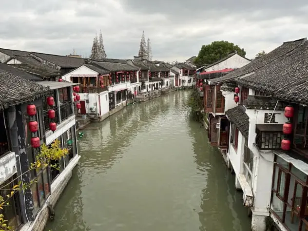 La imagen muestra un pintoresco pueblo acuático tradicional en China, con casas antiguas de techo de teja bordeando un canal. Las farolas rojas decorativas aportan un toque cultural auténtico. Este destino es perfecto para quienes buscan experimentar la tranquilidad de la China histórica, pasear en barca y disfrutar de la arquitectura tradicional. Una joya cultural que combina historia y belleza natural.