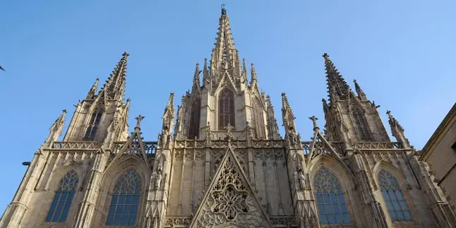 La imagen muestra La imagen muestra la fachada principal de la Catedral de Barcelona, un magnífico ejemplo de arquitectura gótica. Destacan sus imponentes torres y pináculos decorados con detalles ornamentales y gárgolas. En el centro se encuentra el gran ventanal ojival rodeado por tracerías intrincadas, mientras que en la parte superior se erige una alta aguja que domina el cielo despejado. Las ventanas laterales también tienen diseños característicos góticos con arcos apuntados. La piedra, con tonos cálidos y claros, refleja la luz del día, resaltando los detalles arquitectónicos de esta obra histórica.una obra de arte que parece ser de Joan Miró, un famoso artista surrealista conocido por su estilo abstracto y colorido. La obra presenta una serie de formas y líneas abstractas que se entrelazan y se superponen en un fondo de colores suaves, predominantemente tonos de azul y rosa.  Las formas en la pintura incluyen curvas, espirales, puntos y figuras geométricas, muchas de las cuales están coloreadas en negro, rojo, y azul. Hay varios elementos que parecen tener ojos y otras características faciales, lo que es típico del estilo de Miró. Las líneas negras finas conectan varias de estas formas, creando una sensación de movimiento y flujo a lo largo de la obra.  El fondo de la pintura está texturizado con un efecto de pulverización o salpicaduras, que añade profundidad y complejidad al conjunto. La composición general es dinámica y llena de energía, con un equilibrio entre los colores vibrantes y las formas orgánicas y geométricas.  Esta descripción resalta el estilo característico de Joan Miró, conocido por sus representaciones abstractas y su uso audaz del color.