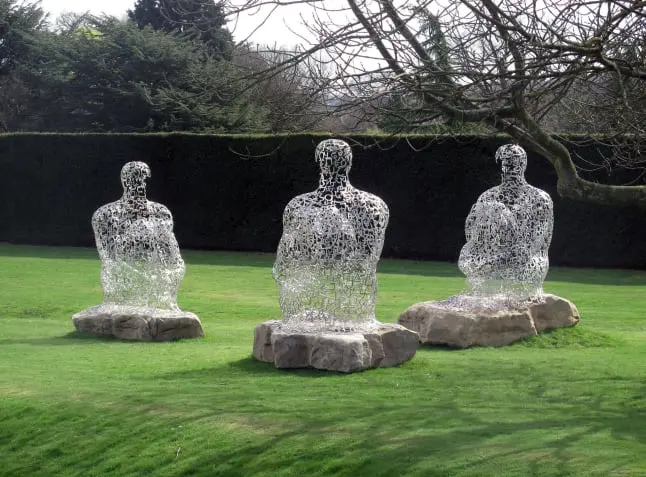 La imagen muestra tres esculturas abstractas hechas de letras metálicas entrelazadas, representando figuras humanas sentadas sobre rocas en un entorno al aire libre. Estas obras, posiblemente del artista Jaume Plensa, exploran el lenguaje, la comunicación y la conexión entre las personas. Las letras que componen las figuras parecen flotar, creando un efecto etéreo que contrasta con el entorno natural del césped verde y los árboles. Las esculturas invitan a la reflexión sobre la interacción entre la humanidad, el arte y la naturaleza, generando un espacio contemplativo en este paisaje.