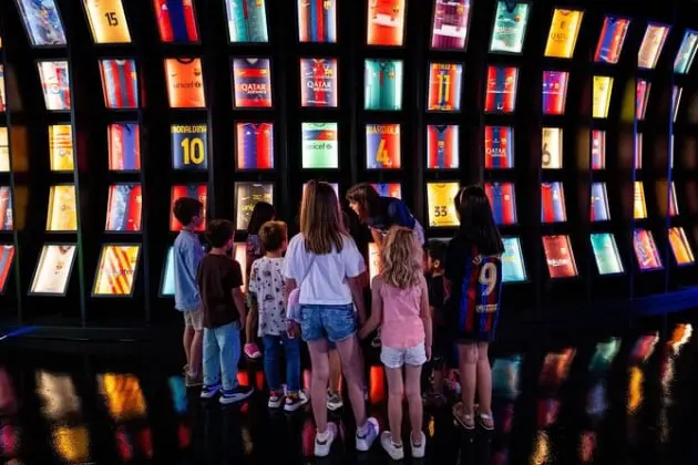 Un grupo de niños observa atentamente una impresionante colección de camisetas históricas del FC Barcelona, expuestas en un museo interactivo. Las vitrinas iluminadas muestran diseños icónicos y números de leyendas como Ronaldinho y Messi. El reflejo en el suelo brillante añade un toque moderno, mientras la escena refleja la pasión y el legado del fútbol en las nuevas generaciones.