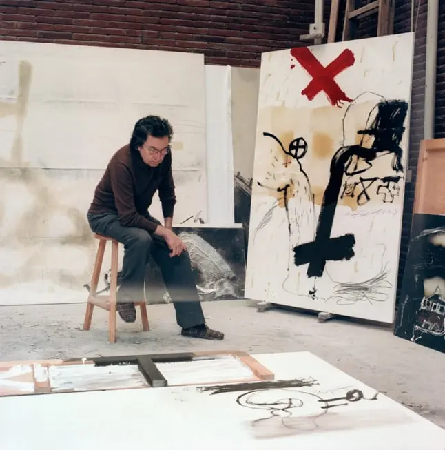 La imagen muestra a un hombre en un estudio artístico, posiblemente el pintor y escultor Antoni Tàpies, conocido por su obra abstracta y expresionista. Está sentado en un taburete, con una postura reflexiva, observando algunas de sus obras en proceso. En el fondo y alrededor, se ven lienzos de gran formato con trazos abstractos, símbolos y texturas, característicos de su estilo. Algunos elementos, como la gran cruz negra y los colores terrosos, reflejan su interés por el simbolismo y las técnicas mixtas. El entorno transmite una atmósfera de creatividad, introspección y experimentación artística.