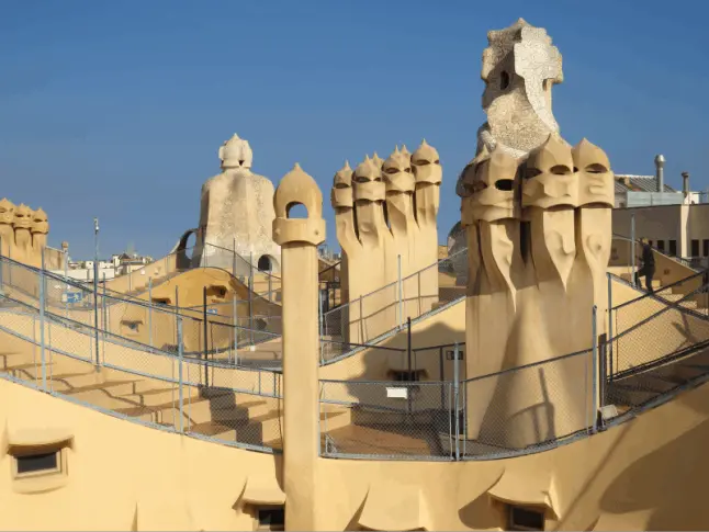 La imagen muestra la terraza de La Pedrera (Casa Milà), una de las obras maestras del arquitecto Antoni Gaudí ubicada en Barcelona. La terraza es conocida por sus emblemáticas chimeneas escultóricas, que combinan funcionalidad y diseño artístico. Estas estructuras tienen formas orgánicas y abstractas, con texturas y curvas que evocan elementos naturales, como máscaras o figuras de guerreros. El acabado de las chimeneas está hecho con piedra, cerámica y otros materiales que reflejan la innovación de Gaudí. El diseño fluido de la terraza, junto con las formas sinuosas de las barandillas y las escaleras, crea un espacio único que mezcla arte, arquitectura y naturaleza, característico del modernismo catalán.