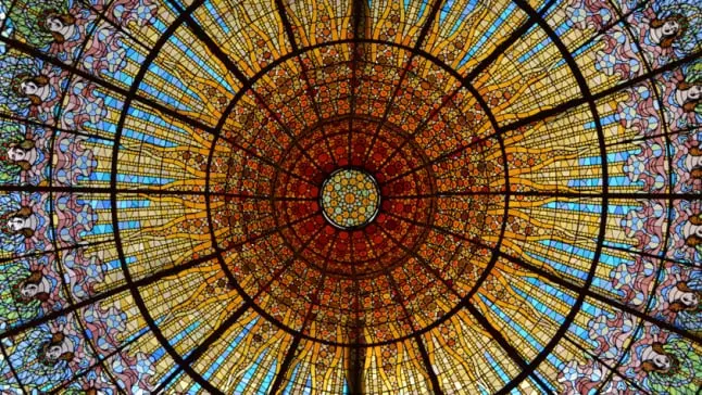 La imagen muestra el espectacular tragaluz del Palau de la Música Catalana en Barcelona, una joya del modernismo catalán. Este vitral circular, diseñado por Antoni Rigalt, representa un gran sol invertido rodeado por tonalidades cálidas, como amarillos y naranjas, que irradian hacia el azul del cielo. El diseño integra detalles florales y figuras femeninas alegóricas, simbolizando la música y la naturaleza. La composición, realizada con intrincadas piezas de vidrio, destaca por su belleza y luminosidad, convirtiéndose en el elemento central que llena de luz y color el auditorio del Palau. Es un ejemplo icónico de artesanía y creatividad arquitectónica.