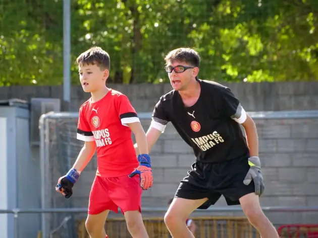 Un joven jugador con uniforme rojo del Campus Girona FC y guantes de portero se concentra en la acción, mientras un entrenador con camiseta negra le guía activamente. Ambos muestran determinación en el campo, reflejando el esfuerzo y el aprendizaje en un ambiente deportivo. Al fondo, el sol ilumina la escena con un entorno natural y competitivo.