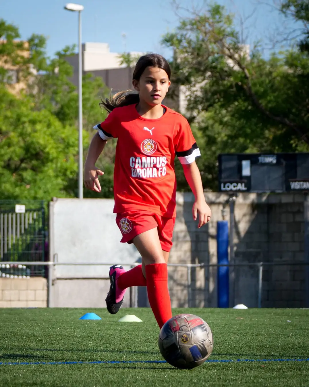 Niña futbolista entrenando en el Campus Girona FC, con camiseta roja y balón en juego. Imagen inspiradora que refleja deporte, esfuerzo y desarrollo juvenil. Ideal para contenido sobre fútbol femenino, campus deportivos, formación infantil y actividades al aire libre para niñas apasionadas por el deporte y el trabajo en equipo.