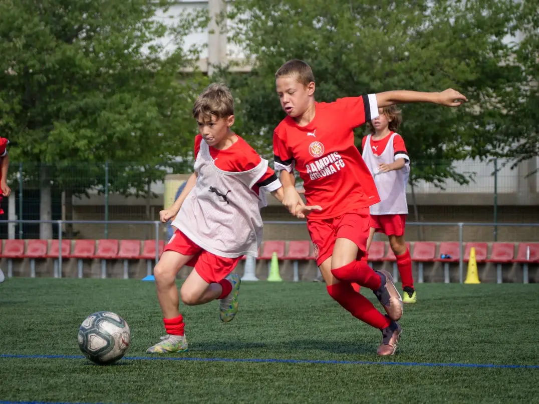 Dos jóvenes futbolistas compiten por el balón en un campo de fútbol durante un entrenamiento en el campus Girona FC. Imagen dinámica que refleja esfuerzo, deporte y desarrollo juvenil. Perfecta para contenido sobre formación deportiva, fútbol base, competiciones infantiles y actividades en equipo para niños y adolescentes.