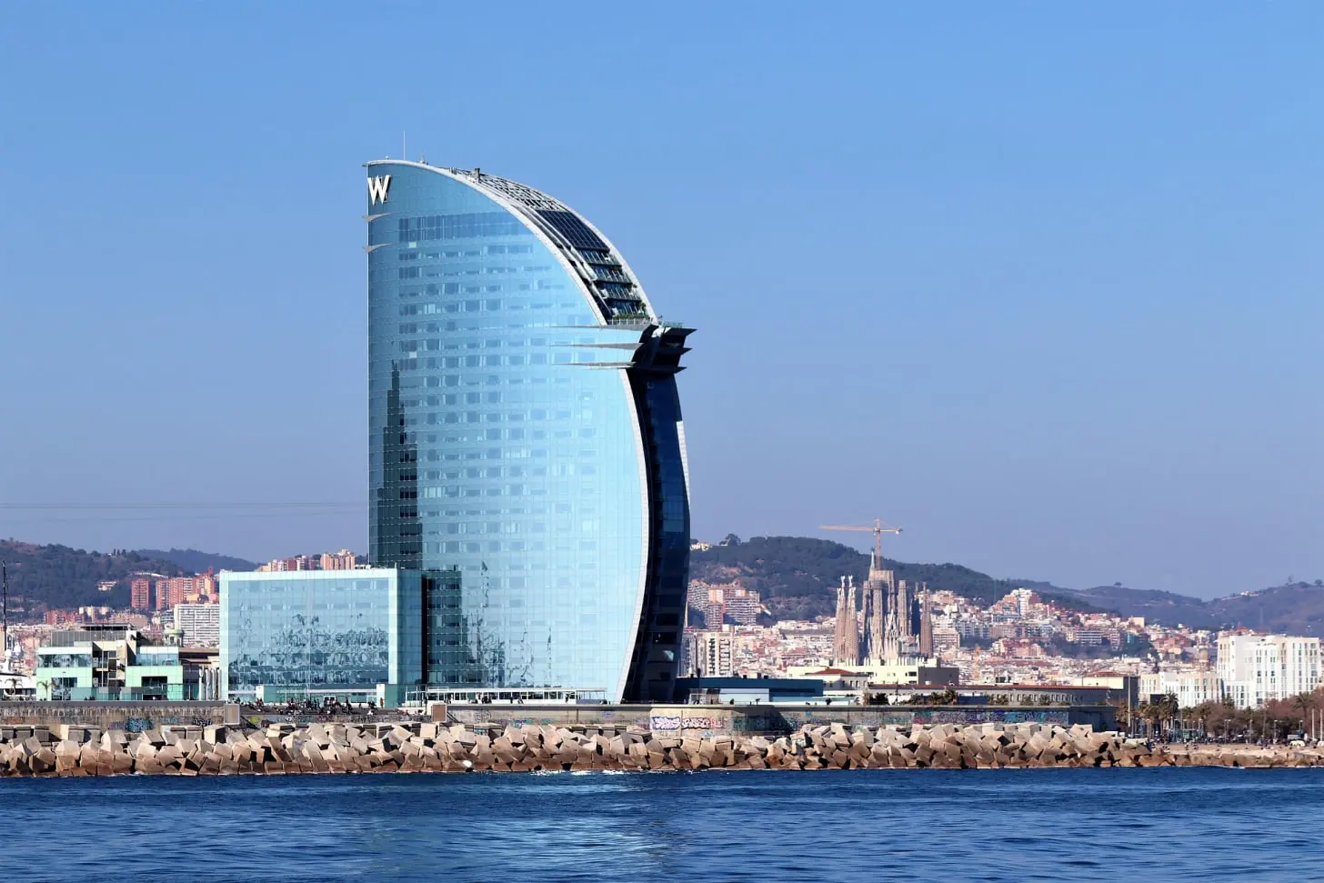 La imagen muestra el famoso Hotel W Barcelona, también conocido como "Hotel Vela" debido a su forma característica de vela náutica. El edificio tiene una estructura moderna, con una fachada predominantemente de cristal que refleja el cielo y el mar, lo que acentúa su integración con el entorno marítimo. Está situado en la Barceloneta, justo al borde del mar Mediterráneo, ofreciendo vistas espectaculares tanto del mar como de la ciudad.  En el horizonte, detrás del hotel, se pueden apreciar partes de Barcelona, con edificios icónicos como la Sagrada Familia, que resalta entre las demás construcciones. El cielo está despejado, lo que sugiere un día soleado, típico del clima mediterráneo de la ciudad. La costa rocosa y el agua en primer plano completan esta imagen de uno de los lugares más emblemáticos del litoral barcelonés.  Este hotel es un referente arquitectónico moderno y una de las construcciones más reconocibles de la ciudad, especialmente por su ubicación privilegiada junto al mar.