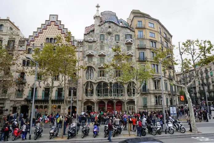 La imagen muestra una de las joyas arquitectónicas más emblemáticas de Barcelona, la Casa Batlló, diseñada por el célebre arquitecto Antoni Gaudí. Este edificio modernista está ubicado en el Paseo de Gracia, una de las avenidas más importantes y elegantes de la ciudad.  La fachada de la Casa Batlló destaca por su diseño orgánico, con formas sinuosas y ventanas irregulares que parecen imitar las ondas del mar. El techo está recubierto de baldosas cerámicas que asemejan las escamas de un dragón, un motivo muy recurrente en las obras de Gaudí, simbolizando la leyenda de Sant Jordi, el patrón de Cataluña. Las barandillas de los balcones recuerdan a máscaras o huesos, lo que otorga a la fachada un aspecto misterioso y naturalista.  A ambos lados de la Casa Batlló, se observan otros edificios igualmente impresionantes, como la Casa Amatller, con su diseño neogótico a la izquierda, reconocible por su forma de escalera invertida en la parte superior. A lo largo de la calle, se puede ver un gran número de personas, probablemente turistas que visitan esta famosa zona. Varias motos están aparcadas en la acera, algo común en las calles céntricas de Barcelona.  El entorno también incluye árboles en primera línea, que indican que probablemente sea otoño debido al aspecto de las hojas. En resumen, la imagen captura un día típico en una de las áreas más turísticas de Barcelona, con una concentración de obras maestras del modernismo catalán.