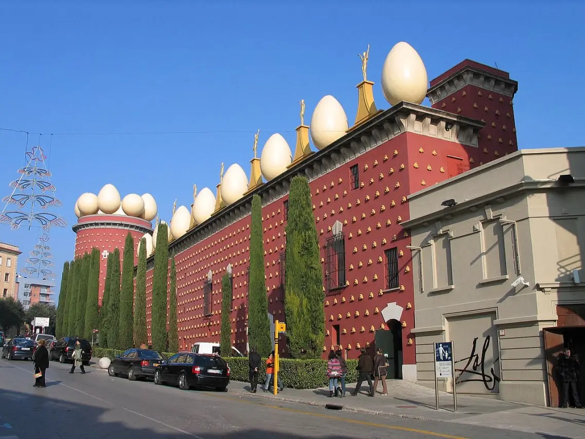 La imagen muestra el exterior del Museo Dalí, ubicado en Figueres, España. Este edificio es conocido por su arquitectura única y surrealista, que refleja el estilo del famoso artista Salvador Dalí.  El museo tiene una fachada de color rojo oscuro adornada con una serie de figuras doradas pequeñas que sobresalen de las paredes. En la parte superior del edificio hay grandes huevos blancos colocados a intervalos regulares, lo que le da un aspecto distintivo e inusual. Estos elementos son característicos del estilo excéntrico y creativo de Dalí.  También se pueden ver árboles altos y delgados, probablemente cipreses, alineados frente al museo, añadiendo un elemento de verdor al entorno urbano. A lo largo de la calle, hay varias personas caminando y coches estacionados, lo que sugiere que es una zona concurrida y popular entre los visitantes.  En la esquina derecha de la imagen, hay una sección del edificio con una firma grande de Salvador Dalí, que refuerza la identidad del museo. El cielo está despejado y azul, lo que indica que es un día soleado.  En conjunto, la imagen captura la esencia surrealista del Museo Dalí, con sus elementos arquitectónicos distintivos y su ambiente artístico vibrante.