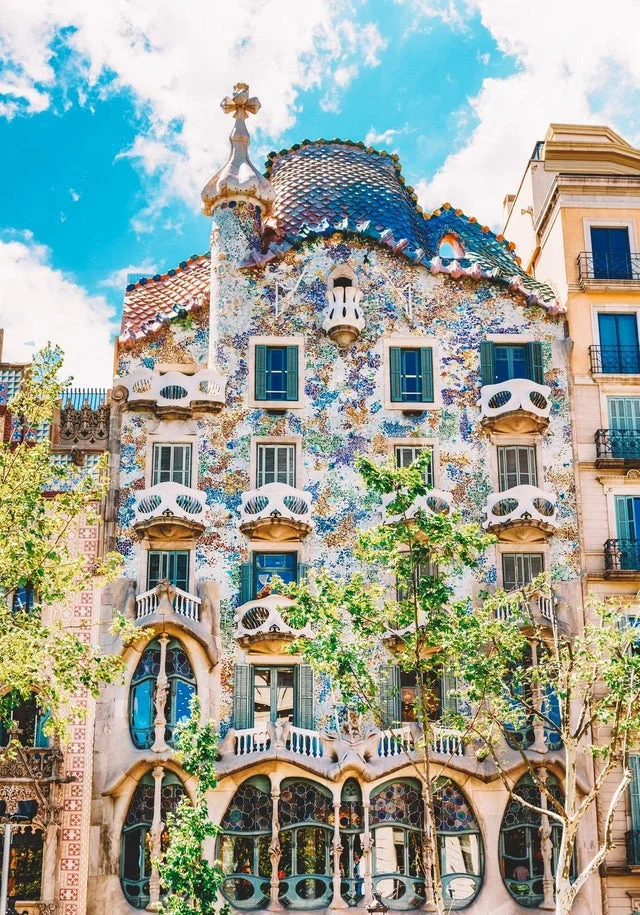 La imagen muestra la Casa Batlló, una de las obras más emblemáticas de Antoni Gaudí ubicada en el Paseo de Gracia de Barcelona. Su fachada destaca por sus formas onduladas, balcones que evocan máscaras y un uso vibrante de mosaicos de cerámica de colores (trencadís), que reflejan luz y aportan un dinamismo único. El tejado, con su forma de lomo de dragón cubierto de escamas, y la torre coronada con una cruz de cuatro brazos, añaden un simbolismo característico del estilo modernista catalán. Este edificio es un ícono de creatividad y arquitectura innovadora, atrayendo a visitantes de todo el mundo.