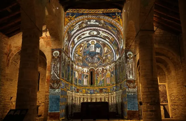 La imagen muestra el ábside de la iglesia románica de Sant Climent de Taüll, en el Valle de Boí, famosa por sus frescos del siglo XII. En el centro, destaca el "Pantocrátor", una representación de Cristo en majestad rodeado por una mandorla y símbolos de los evangelistas. El arte románico, con sus figuras estilizadas y colores vibrantes, resalta la solemnidad espiritual del lugar. Estos frescos son un icono del arte medieval catalán y se encuentran entre las obras más representativas del románico europeo, siendo un referente cultural y artístico de gran valor histórico.