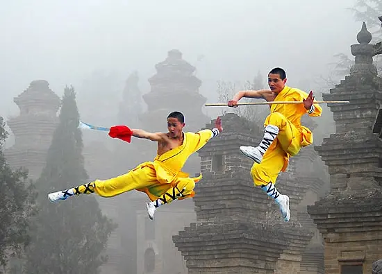 La imagen muestra a dos monjes Shaolin en medio de una demostración acrobática de artes marciales. Vestidos con túnicas amarillas tradicionales, realizan saltos coordinados en el aire, uno portando un bastón y el otro una bandera. De fondo, se observan estructuras antiguas rodeadas por niebla, probablemente en el templo Shaolin, destacando la conexión entre disciplina, cultura y espiritualidad.