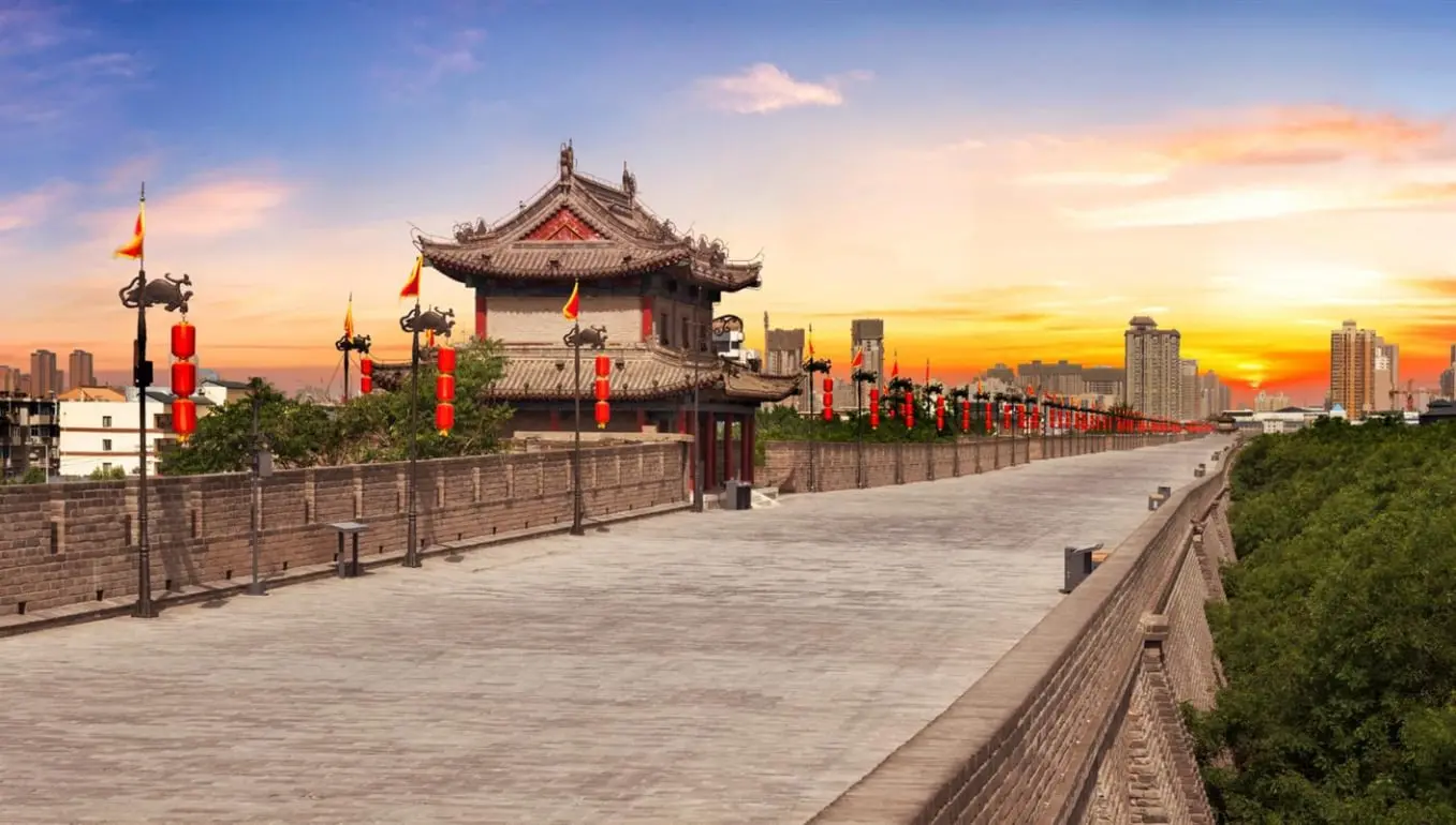 Esta imagen muestra la majestuosa Muralla de Xi'an, uno de los mejor conservados ejemplos de arquitectura militar antigua en China. La escena captura un atardecer impresionante sobre la muralla, con los característicos farolillos rojos y el pabellón tradicional que añaden un toque cultural distintivo. Construida durante la dinastía Ming, la Muralla de Xi'an es un testimonio de la ingeniería defensiva china y ofrece vistas panorámicas del contraste entre la arquitectura histórica y los modernos edificios urbanos. Un atractivo turístico imprescindible para quienes visitan Xi'an, la ciudad conocida por sus Guerreros de Terracota.